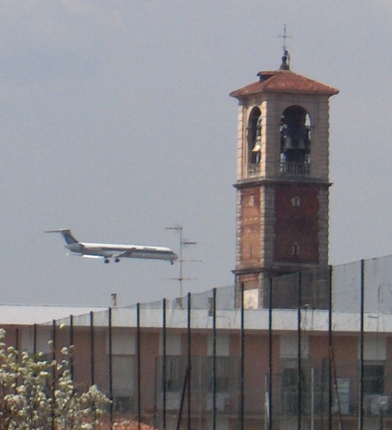 L'antico e il recente: un aereo, in atterraggio presso Malpensa 2000, sfiora il campanile in cui rilucono le campane ripulite. Il tutto  visto da via Antonio da Lonate, dietro l'Oratorio Maschile (foto dell'autore di questo sito)